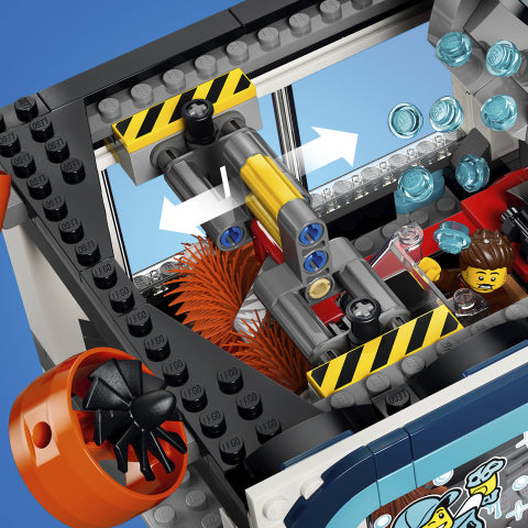 Close-up of a LEGO city construction vehicle or building set interior with gray and black brick frames, yellow and orange mechanical parts, blue translucent blocks, and a small minifigure driver.