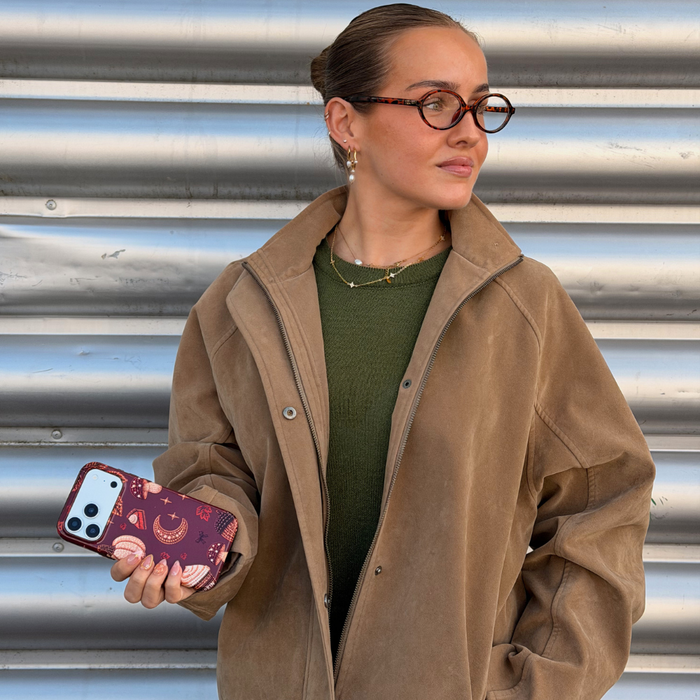 A woman with glasses and her hair in a bun holds a phone with a decorative autumn-themed case featuring illustrations of pumpkins, a crescent moon, and a slice of pumpkin pie.