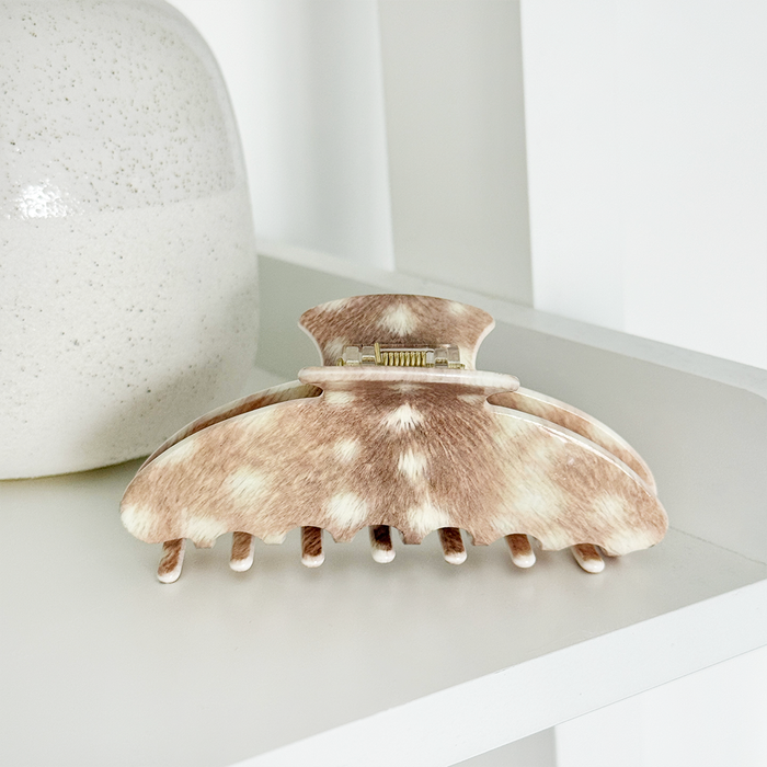 Hair claw clip with a brown and cream mottled animal-print pattern, resting on a white shelf next to a textured ceramic vase.