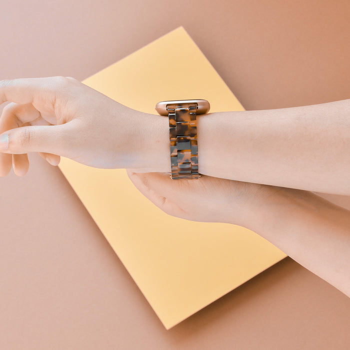 Hand wearing a Coconut Lane Luxe Tortoiseshell Fitbit strap, resting on a yellow notebook against a beige surface.