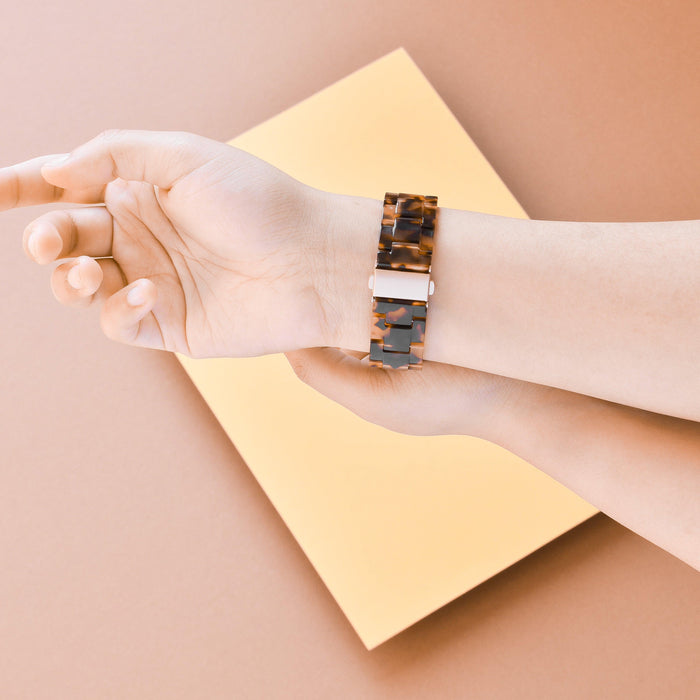 Coconut Lane LUXE Tortoiseshell Fitbit strap on a hand, set against a peach background with a yellow rectangle.
