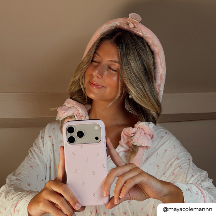 A woman takes a mirror selfie holding a light pink floral phone case and wearing a pink heatless curl set in her hair.