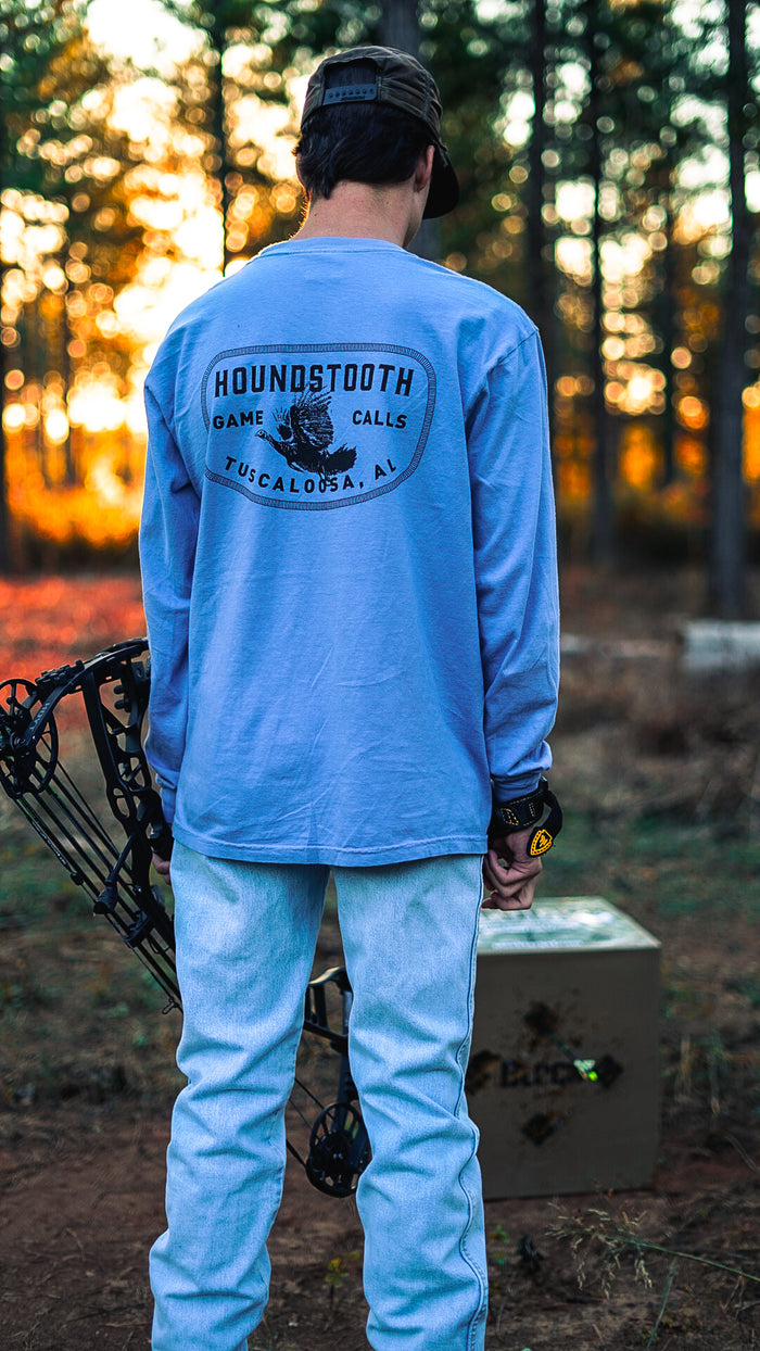 A person wearing a long sleeve blue t-shirt with the text 'HOUNDSTOOTH FLYING LONGBEARD' on the back, holding a bow and arrow, with a blind or shelter in the background.