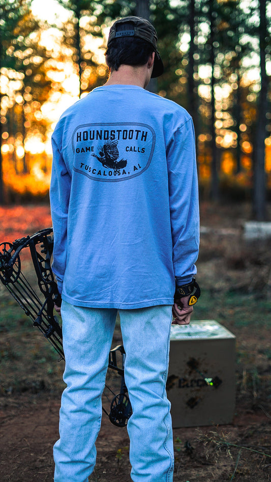 A person wearing a long sleeve blue t-shirt with the text 'HOUNDSTOOTH FLYING LONGBEARD' on the back, holding a bow and arrow, with a blind or shelter in the background.