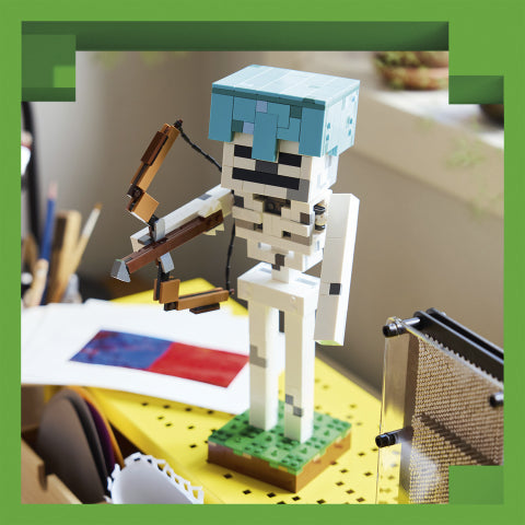 A pixelated toy figure resembling a skeleton or warrior built from block-like pieces, standing on a green base and holding a brown bow. The figure is photographed on a desk with papers and office items in a real indoor setting, so this is Lifestyle rather than Product/Packaging.
