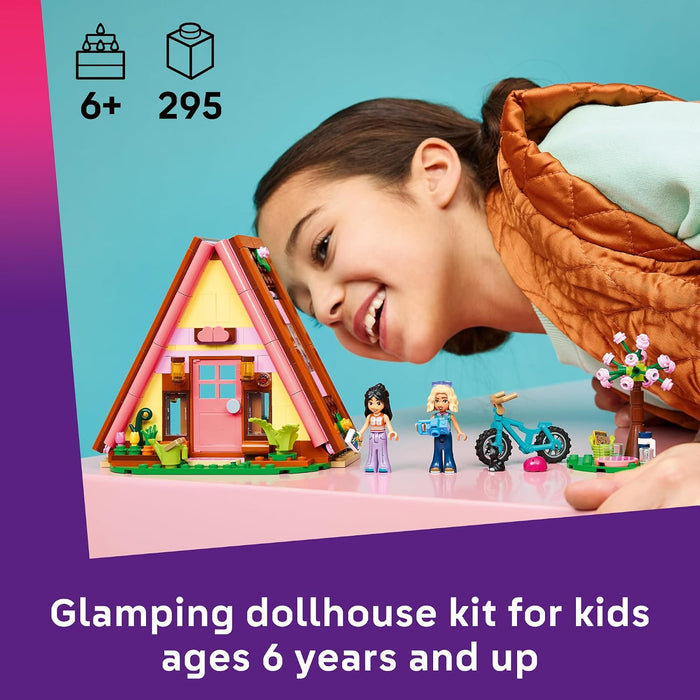 A child smiling beside a colorful glamping dollhouse kit with small figures, a bicycle, and a tree, on a pink tabletop against a turquoise background. Purple text at the bottom reads 'Glamping dollhouse kit for kids ages 6 years and up.