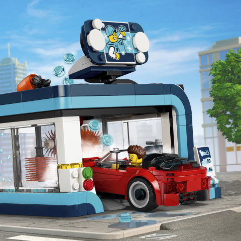 A red brick car with a yellow minifigure driver is parked inside a blue and white toy car wash building, with a large blue and white sign on top and a city street scene in the background.