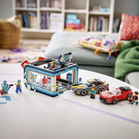 A LEGO City Garage set displayed on a white table in a cozy, blurred living room setting. The set includes a blue and gray garage structure, a yellow and gray sports car, a red sports car, and several LEGO minifigures.