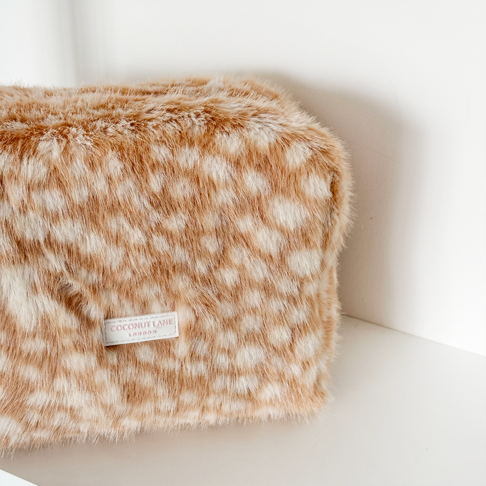A close-up of a fluffy faux-fur cosmetic pouch in a warm tan and cream spotted pattern, with a visible Coconut Lane London label, resting on a white shelf against a white wall.