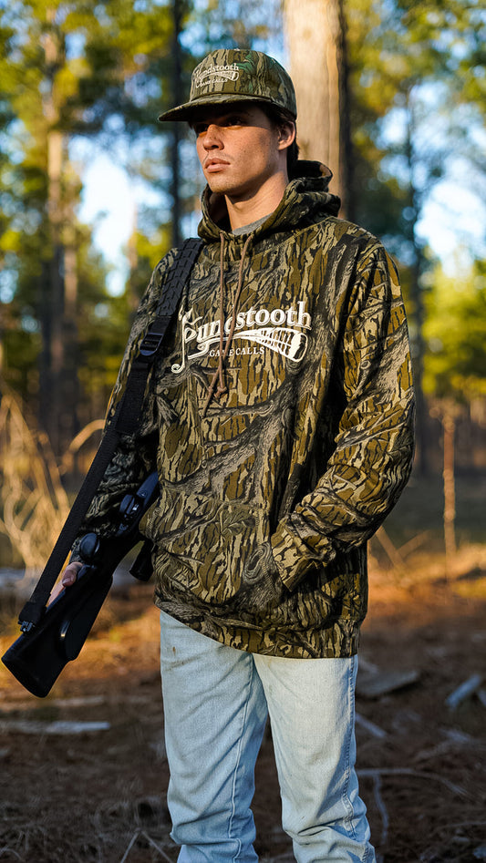 A person wearing a camouflage treestand hoodie with the text 'Mossy Oak' on the front, holding a rifle, and standing in a wooded area.