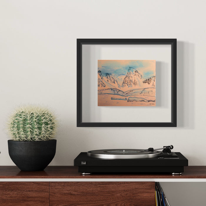The artwork 'Orne Harbour' by Ali Ha depicts a serene mountain landscape in delicate blue and peach tones on warm white paper. The composition features jagged peaks rising against a soft sky, rendered in Micron ink and Caran d'Ache media, displayed in a sleek black frame alongside a cactus plant and record player.