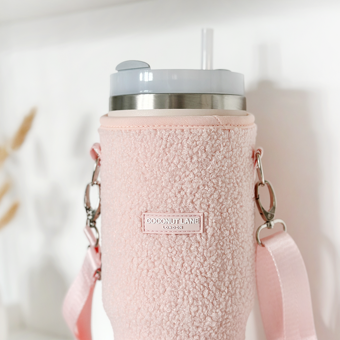 A close-up of a large tumbler with a straw and lid, encased in the Teddy Love-A-Lot Tumbler Carry Case from Coconut Lane. This light pink, textured sherpa sleeve features a label with "Coconut Lane London" and comes with handles attached, making it an adorable accessory for your tumbler. The background is blurred, drawing attention to the carry case.
