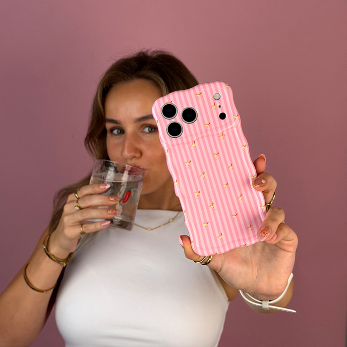 A woman taking a selfie, holding a wavy pink striped phone case with small spicy margarita illustrations, held by a person wearing a white top and black jeans against a solid pink background. She is sipping from a glass with red chillis on it.